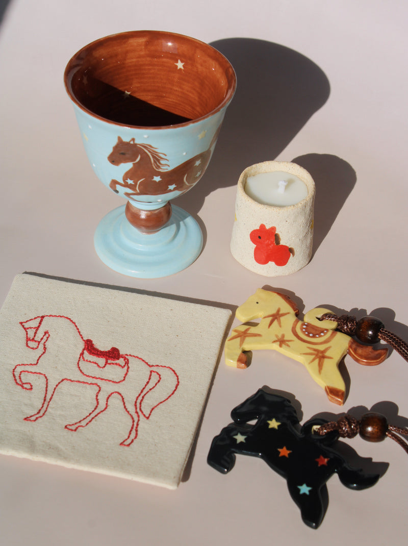 Collection of horse-themed items including a ceramic cup, candle, keychain, and embroidered fabric on a light surface.
