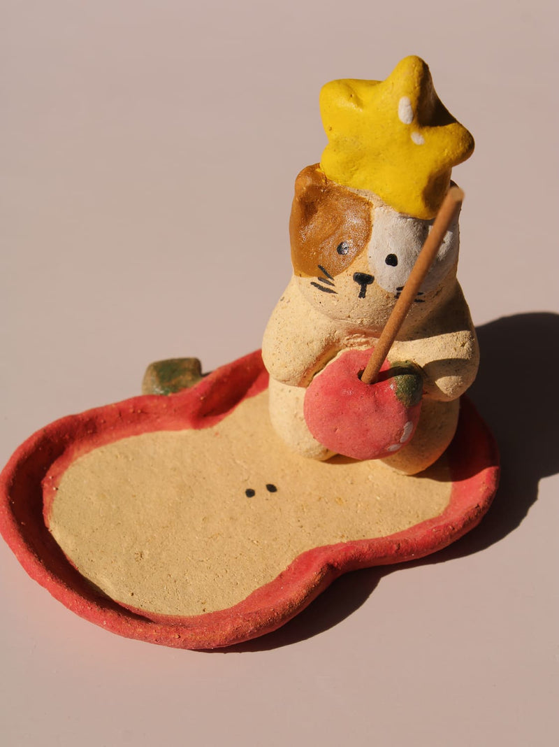 Mochi Ceramic Incense Holder, a handmade cat figurine wearing a yellow star hat and holding a pink apple, paired with an apple-shaped incense tray, on a soft pink background.