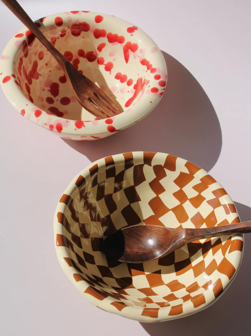 Dotty Ceramic Bowl and Nova Ceramic Bowl side by side with wooden spoons — red dot pattern and brown checkerboard pattern — on a soft pink background.