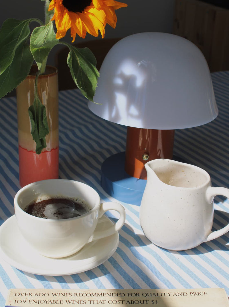 A styled tabletop scene with a white ceramic coffee cup and saucer filled with coffee, a small white creamer pitcher, a mushroom-shaped table lamp, a striped blue-and-white cloth, and a vase with a sunflower.