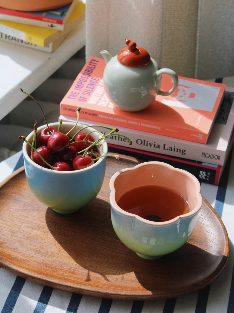 A styled tabletop scene with two pastel ceramic cups on a round wooden tray, one filled with red cherries and the other with amber tea. A small pale green teapot with a red lid sits on stacked books in the background.