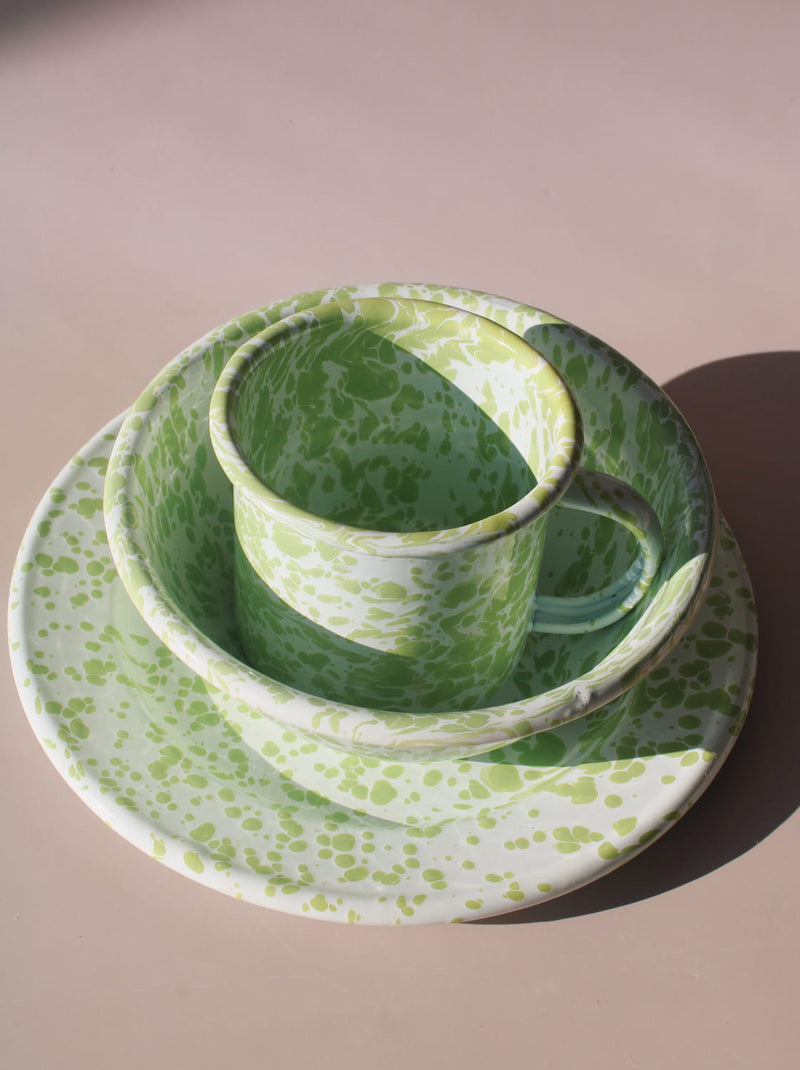 Green splatter enamel tableware set with mug, bowl and plate styled in natural light with wooden spoon.