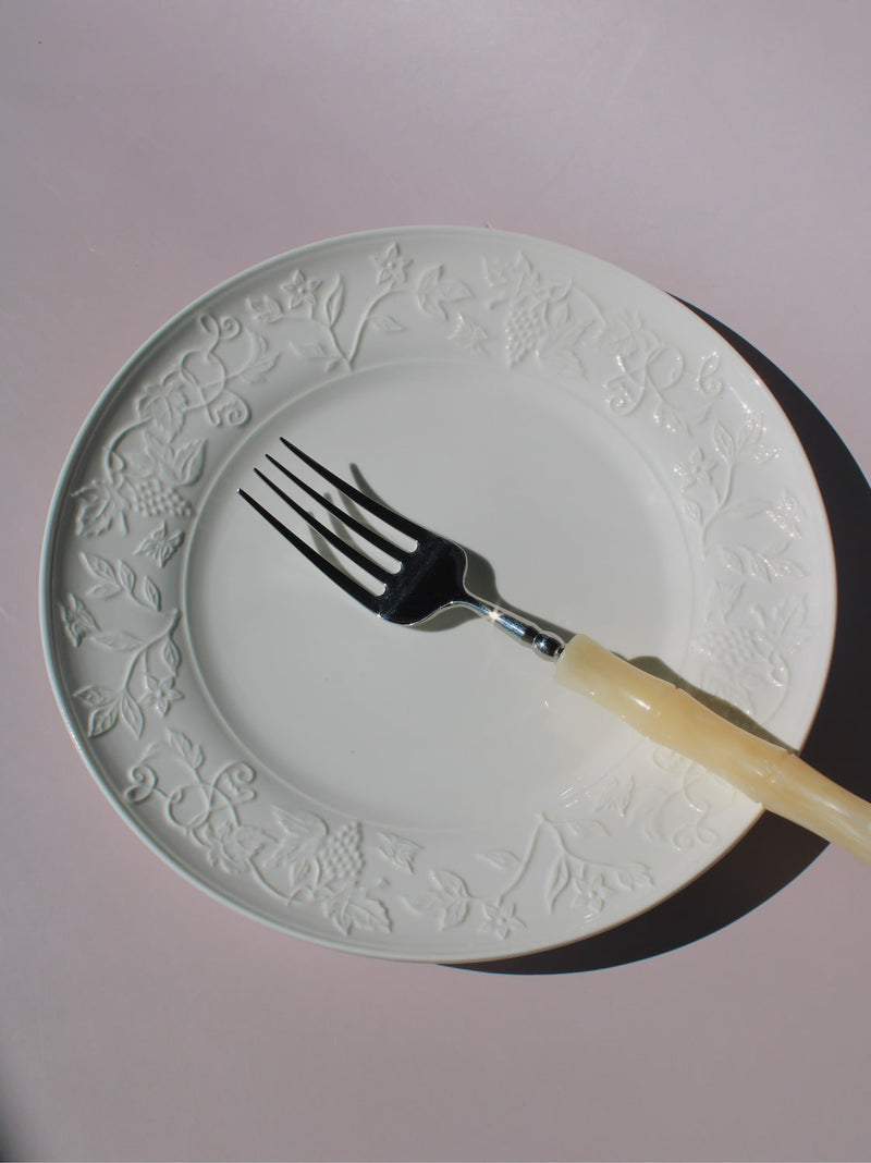 Florentia Ceramic Plate in white with embossed floral and botanical border detail, styled with a silver fork, on a soft pink background.