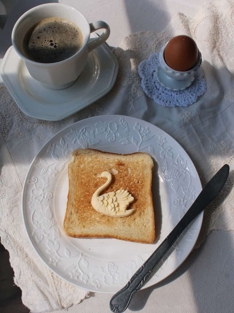A styled breakfast scene with a white plate holding a toasted slice topped with a swan-shaped pastry, alongside a cup of coffee and a hard-boiled egg on a lace cloth.