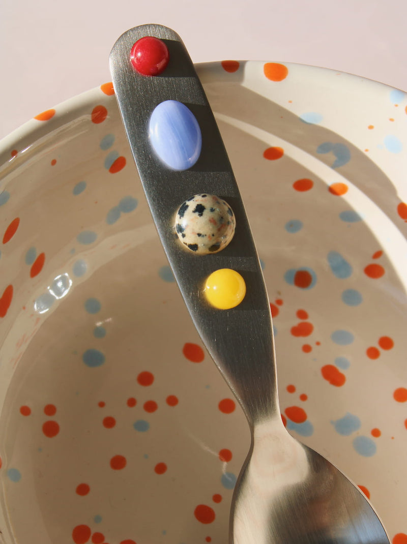 Handmade ceramic speckled bowl with stainless steel dessert spoon featuring colorful bead handle, playful artisan tableware for breakfast or dessert.