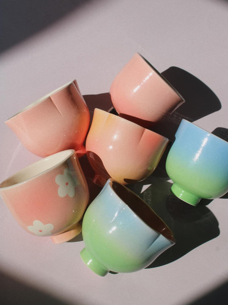handmade ceramic cups from the Yunomi collection in pastel pink, blue and peach ombré glazes, some with scalloped rims and one with a hand-painted flower, arranged on a pink surface in natural light