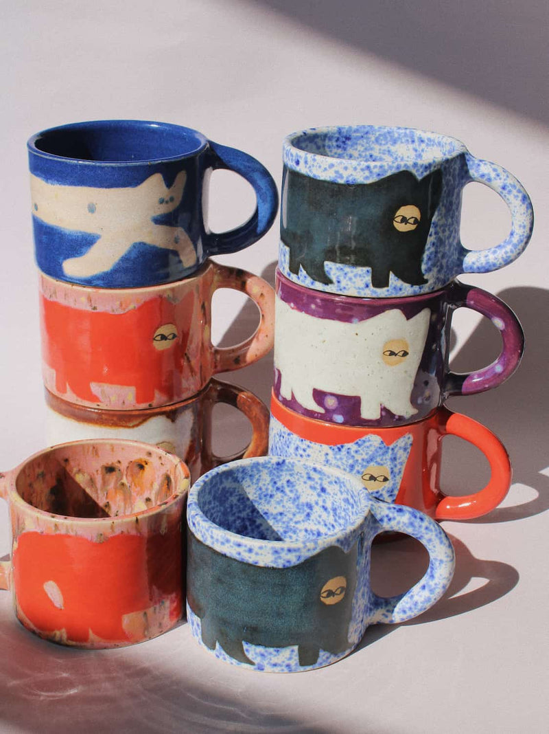 A set of handmade ceramic mugs stacked in two groups, featuring a hand-painted black cat design on speckled blue, orange, purple, and white glazes, with one mug showing a raw, unglazed interior.