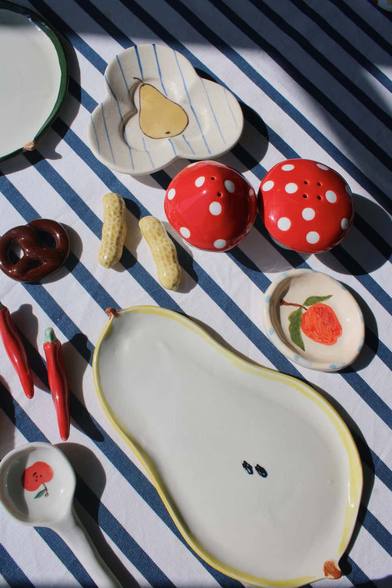 A collection of hand-painted ceramic tableware, including a large pear-shaped plate, red and white polka dot salt and pepper shakers, a strawberry spoon, and other matching dishes, arranged on a blue and white striped fabric under direct sunlight.