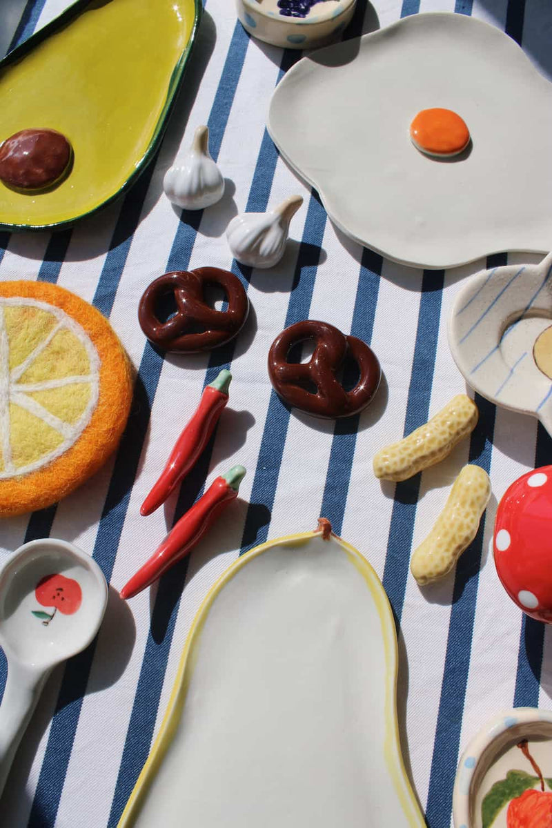 A collection of whimsical, food-themed ceramic plates and small dishes arranged on a blue and white striped tablecloth under bright sunlight. The set includes plates shaped like an avocado half, a fried egg, a lemon slice, pretzels, red chili peppers, peanuts, and a red apple slice, alongside matching spoons and bowls.