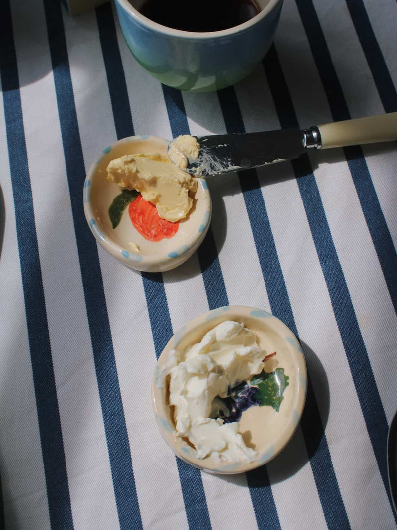 Two small decorative ceramic bowls containing whipped cream and butter, with a knife resting between them on a blue and white striped tablecloth. A ceramic mug is partially visible at the top.