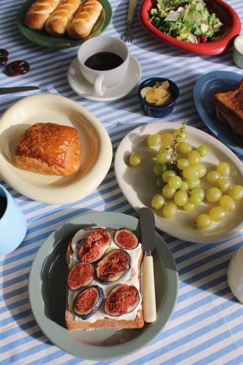 Breakfast table styled with handmade ceramic plates and bowls, featuring pastries, fruit, and coffee on a striped tablecloth