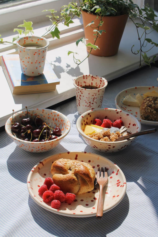 Speckled ceramic mugs, bowls with cherries and fruit, and a plate with a pastry and raspberries, styled on a striped tablecloth by a window.