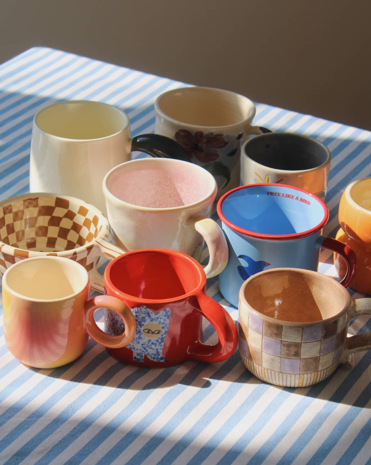 Assorted handmade ceramic mugs in colorful designs and glazes styled on a striped tablecloth for coffee and tea