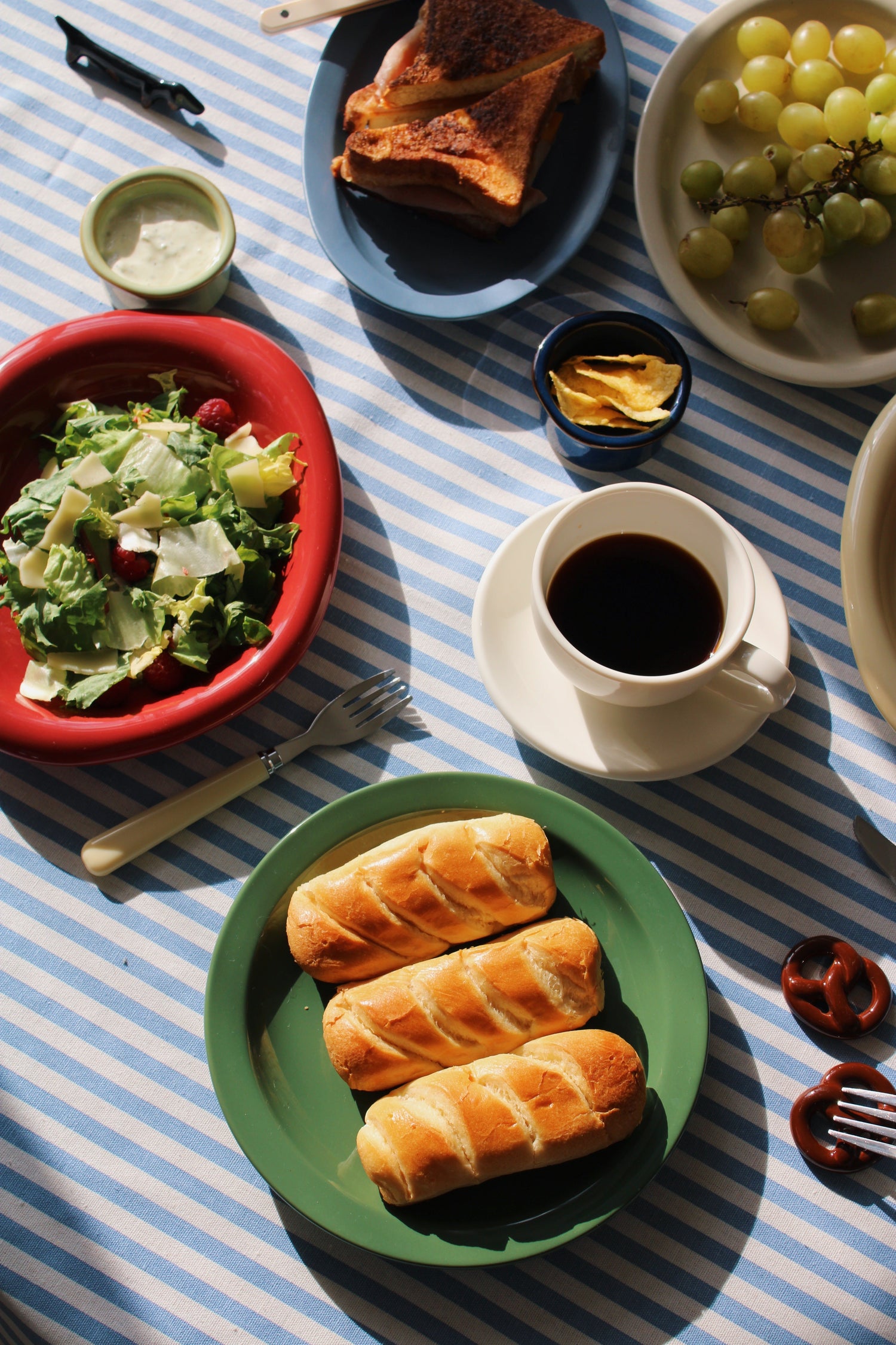 A casual table setting with handmade ceramic plates, coffee, bread, and fresh food styled on a blue striped tablecloth.
