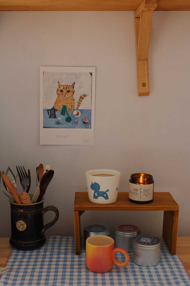 Marcel Cat Art Print – illustrated orange cat art print styled on a wall above a colourful coffee station with ceramic mugs, books and home accessories