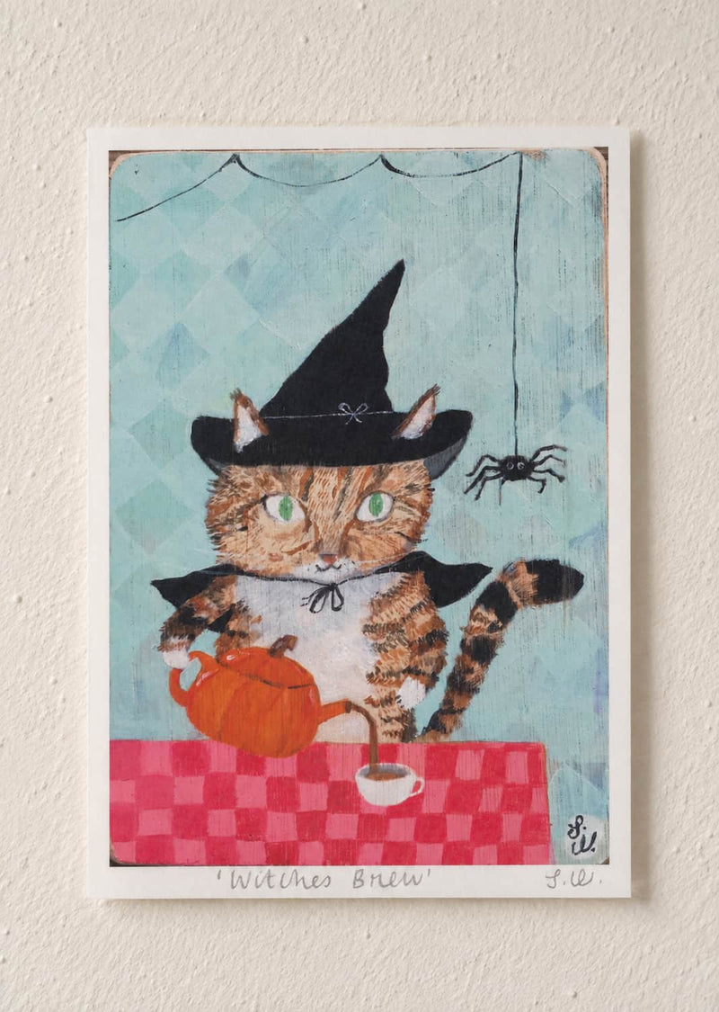 Witches Brew Cat Art Print A whimsical art print titled 'Withes Brew' featuring a tabby cat dressed as a witch holding an orange teapot behind a red checkered table. The print is mounted on a textured off-white wall.