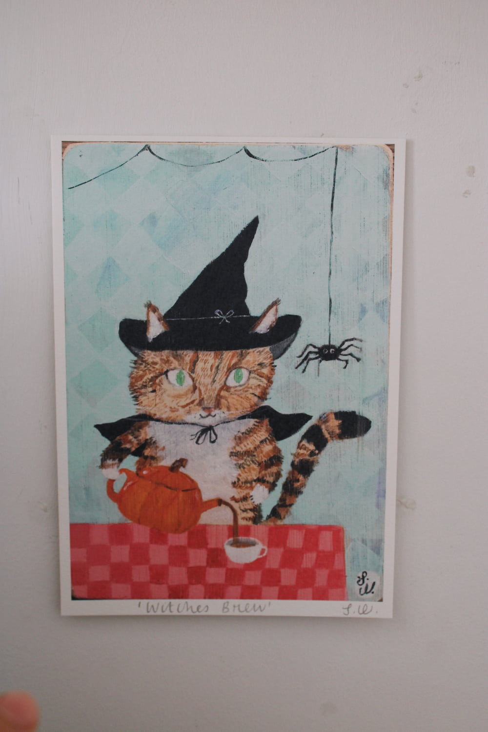 Witches Brew Cat Art Print A whimsical art print titled 'Withes Brew' featuring a tabby cat dressed as a witch holding an orange teapot behind a red checkered table. The print is mounted on a textured off-white wall.