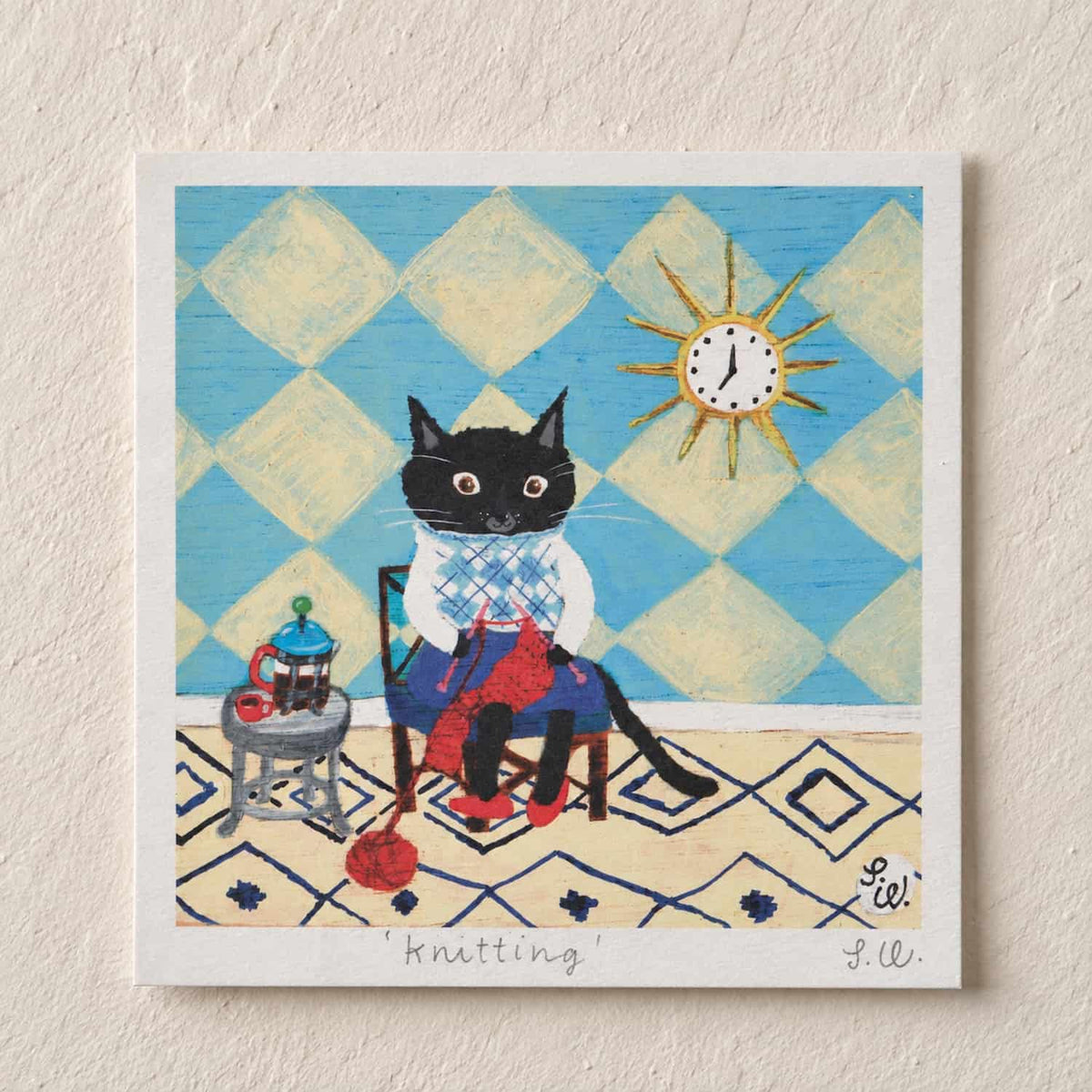 Knitting Cat Art Print featuring a black cat knitting with red yarn in a cozy blue-and-cream room, with a teapot, geometric rug, and sun-shaped clock.