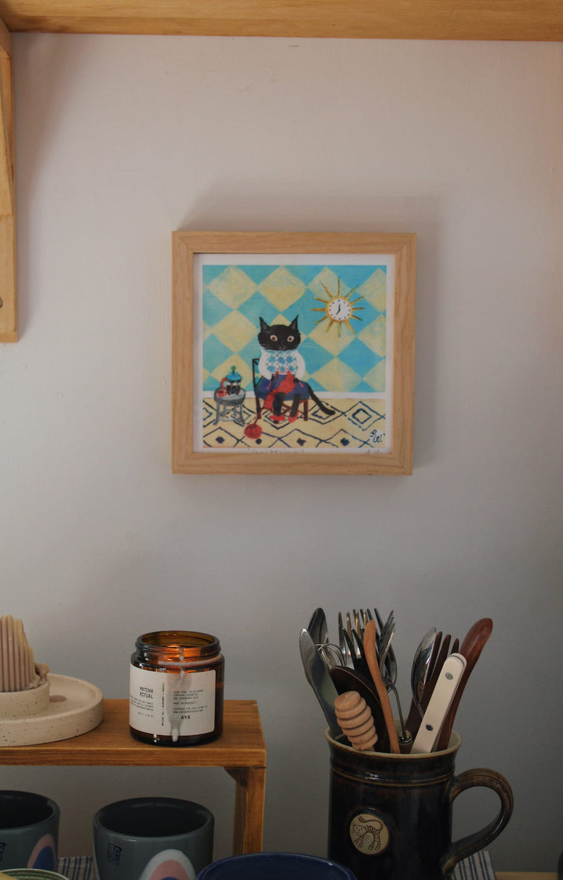 Knitting Cat Art Print – illustrated cat art print styled on a wall above a colourful coffee station with ceramic mugs, books and home decor