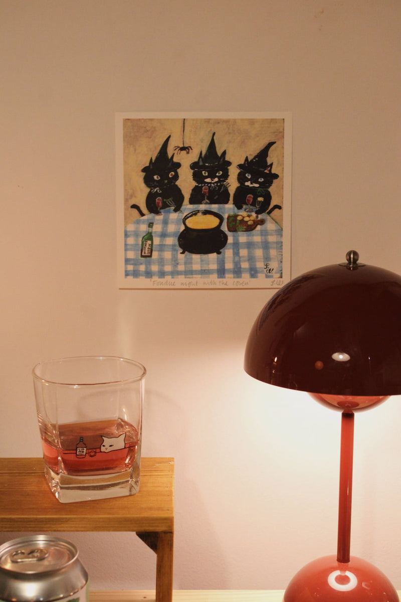 Fondue Night with the Coven Art Print – illustrated art print featuring three black cats dressed as witches around a fondue pot, styled above a wooden side table with a glass and a red table lamp