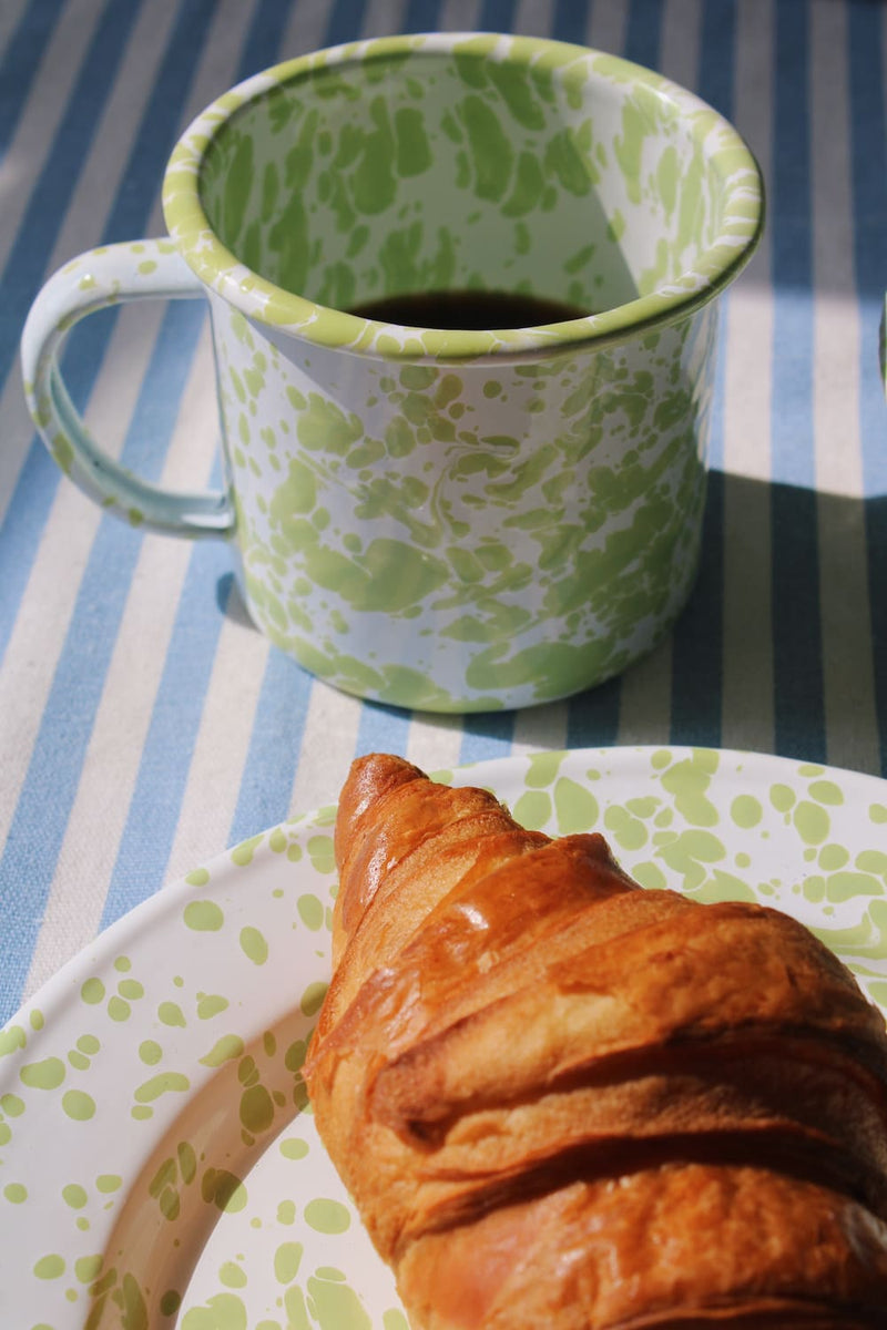 Green patterned clover enamel mug with coffee next to a croissant on a striped tablecloth.