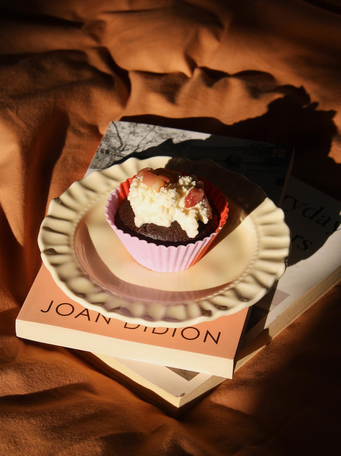 A chocolate cupcake with white frosting and red berries sits on a white scalloped plate, placed on top of a Joan D'ion book against a warm brown draped fabric background.