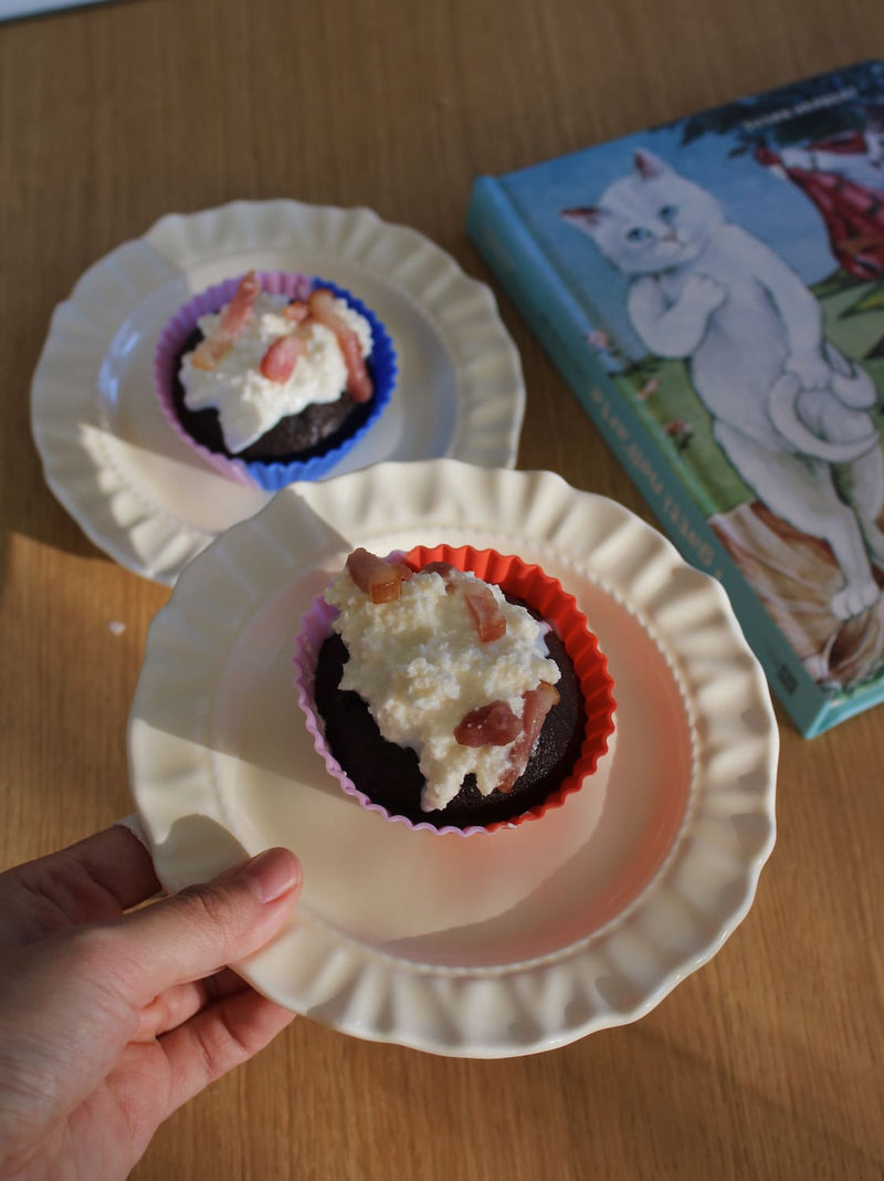 A hand holds a white paper plate with a chocolate cupcake topped with whipped cream and dried fruit pieces in a red liner. Another cupcake on a blue liner sits on a white plate in the background, with a blue book featuring a white cat on a wooden table.