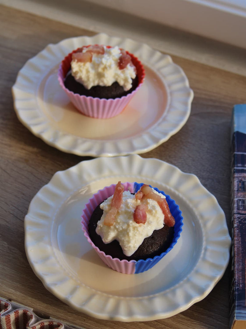 Two small cupcakes topped with whipped cream and fruit pieces, each in a colorful paper liner on a white scalloped plate, set on a wooden surface.