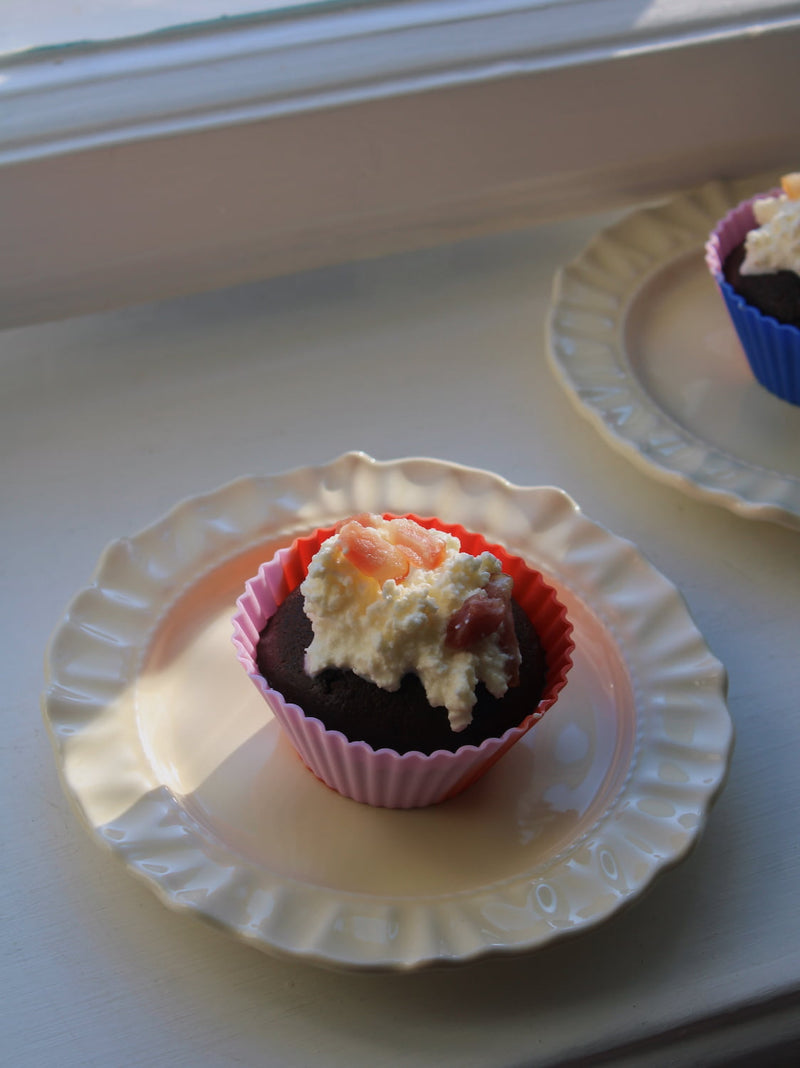 A chocolate cupcake in a pink paper liner with whipped cream and fruit topping, sitting on a white scalloped plate on a windowsill.