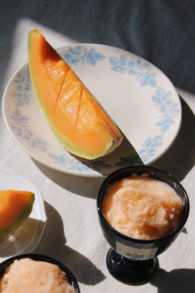 A styled food scene with a sliced cantaloupe melon on a white plate with a blue floral border, alongside two small black cups filled with a pale peach-colored frozen dessert or ice cream on a sunlit white surface.
