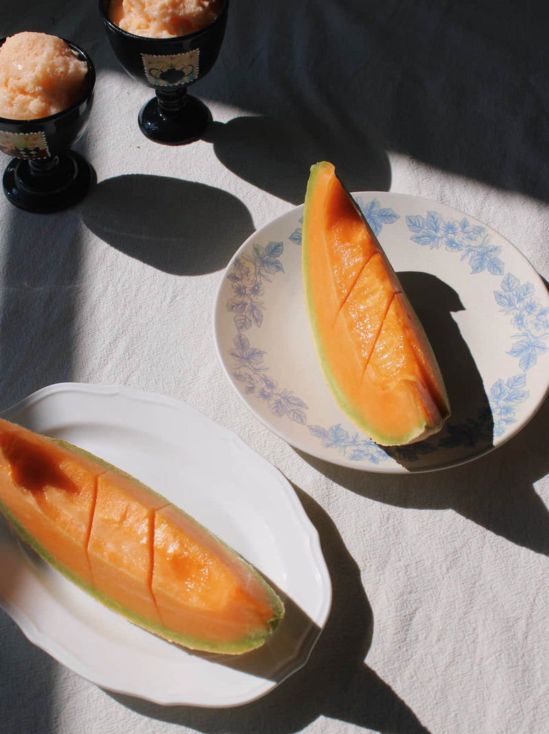 Two slices of orange cantaloupe melon arranged on white plates with blue floral detailing, set on a white fabric surface under sharp natural sunlight with strong shadows.