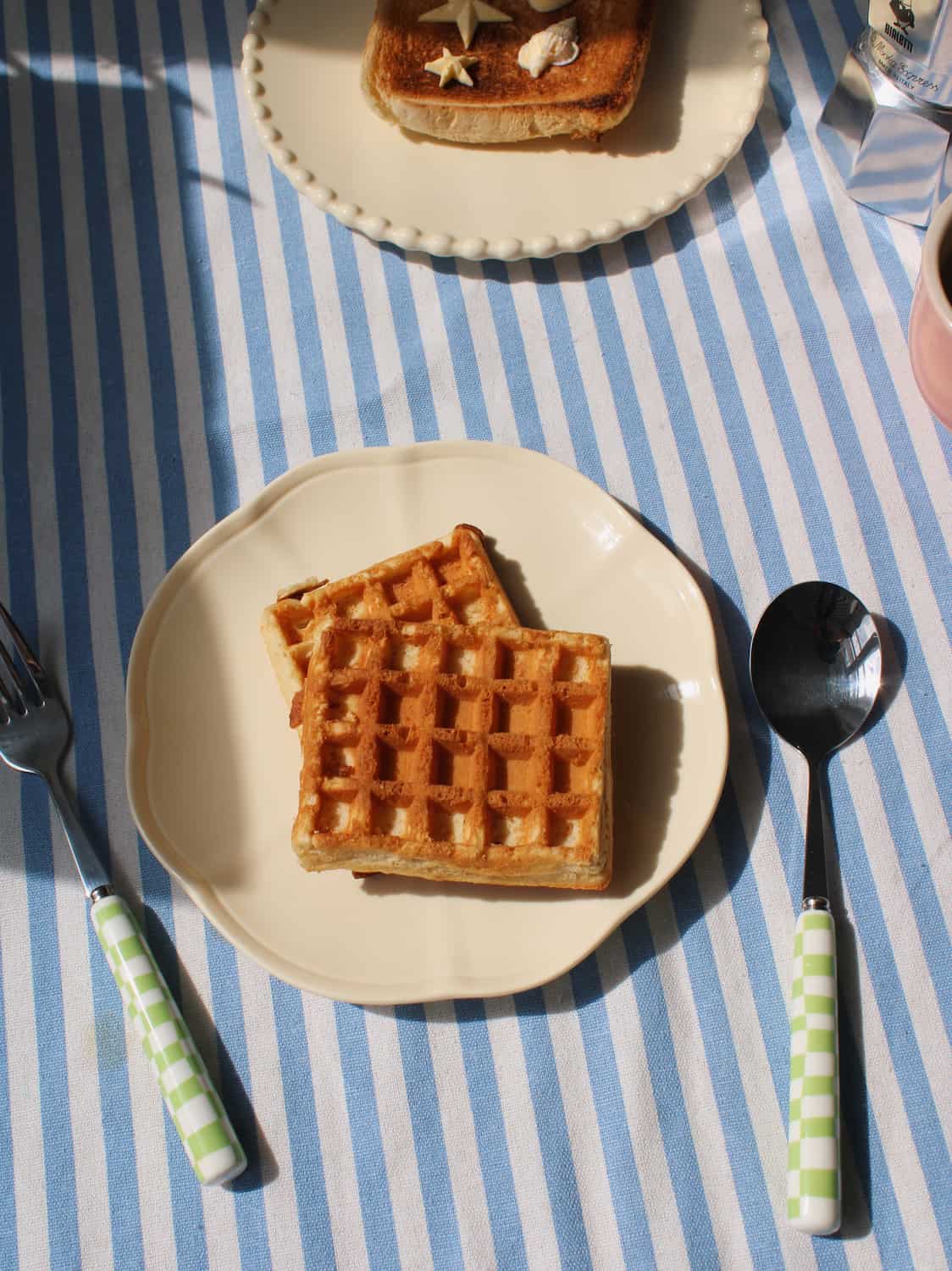 Two square waffles on a cream-colored plate on a blue and white striped tablecloth, with a spoon and fork featuring green-and-white checkered handles beside it.