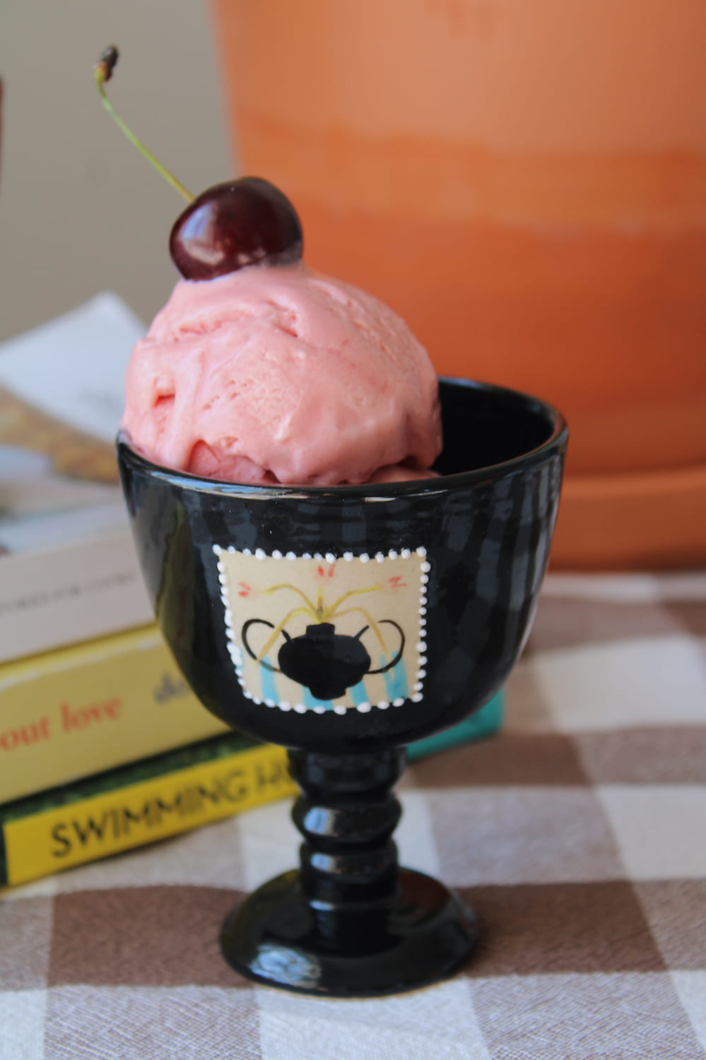 Two Ophelia Ceramic Goblets in glossy black with hand-painted stamp motifs, filled with strawberry and mango ice cream topped with a cherry and raspberry, styled on a stack of books by a sunny window.