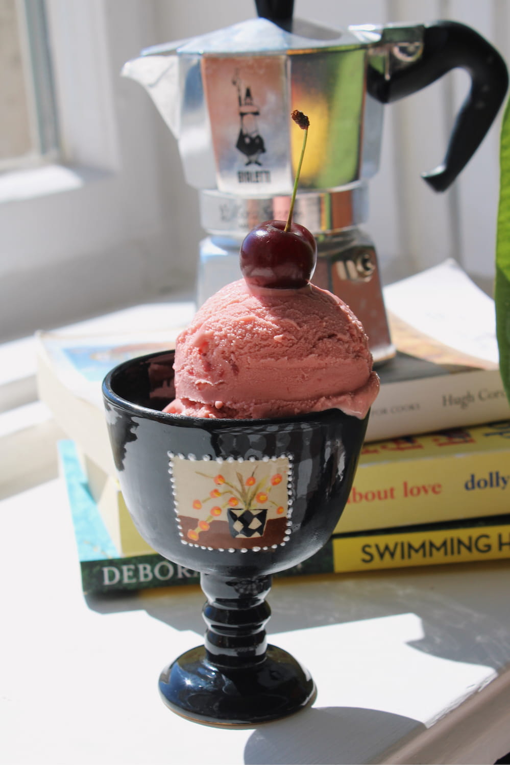 Two Ophelia Ceramic Goblets in glossy black with hand-painted stamp motifs, filled with strawberry and mango ice cream topped with a cherry and raspberry, styled on a stack of books by a sunny window.