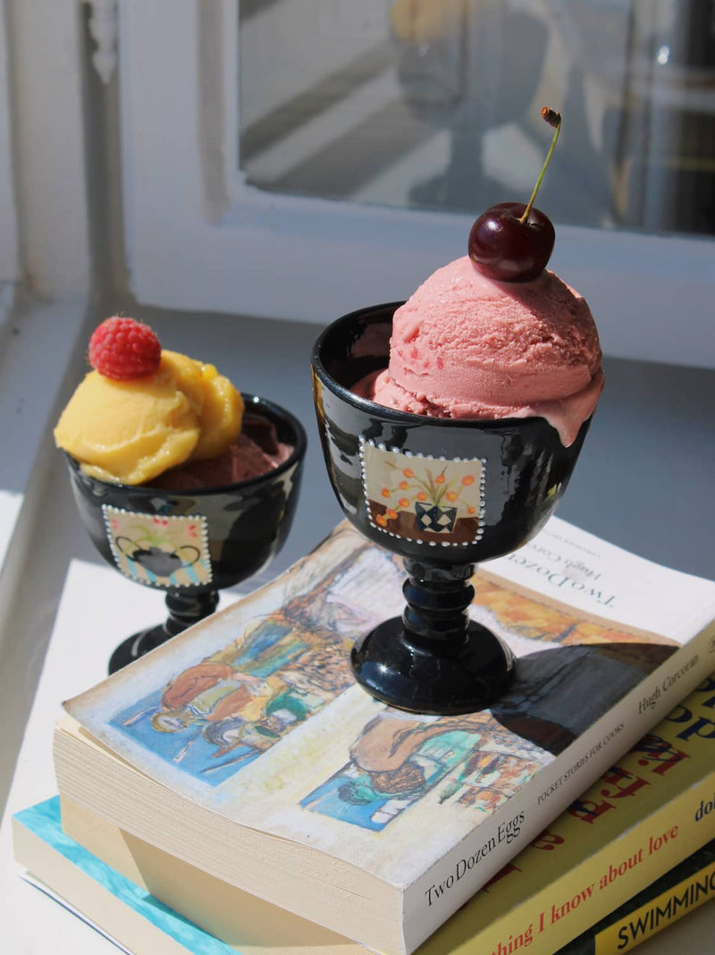 Two Ophelia Ceramic Goblets in glossy black with hand-painted stamp motifs, filled with strawberry and mango ice cream topped with a cherry and raspberry, styled on a stack of books by a sunny window.