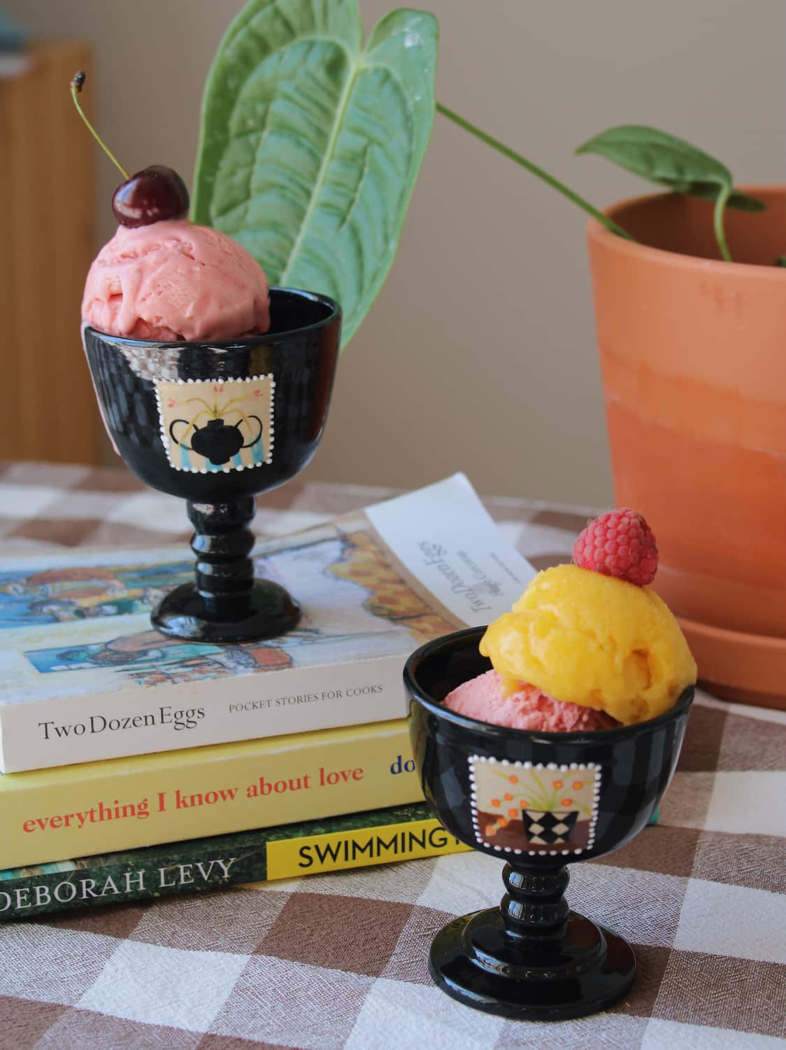 Two Ophelia Ceramic Goblets in glossy black with hand-painted stamp motifs, filled with strawberry and mango ice cream topped with a cherry and raspberry, styled on a stack of books by a sunny window.