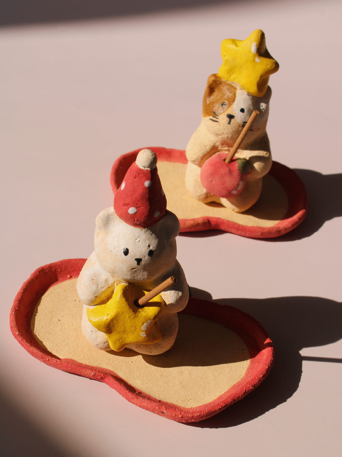 Teddy Ceramic Incense Holder and Mochi Ceramic Incense Holder, two handmade animal figurines with star and apple accents on matching apple-shaped incense trays, on a soft pink background.
