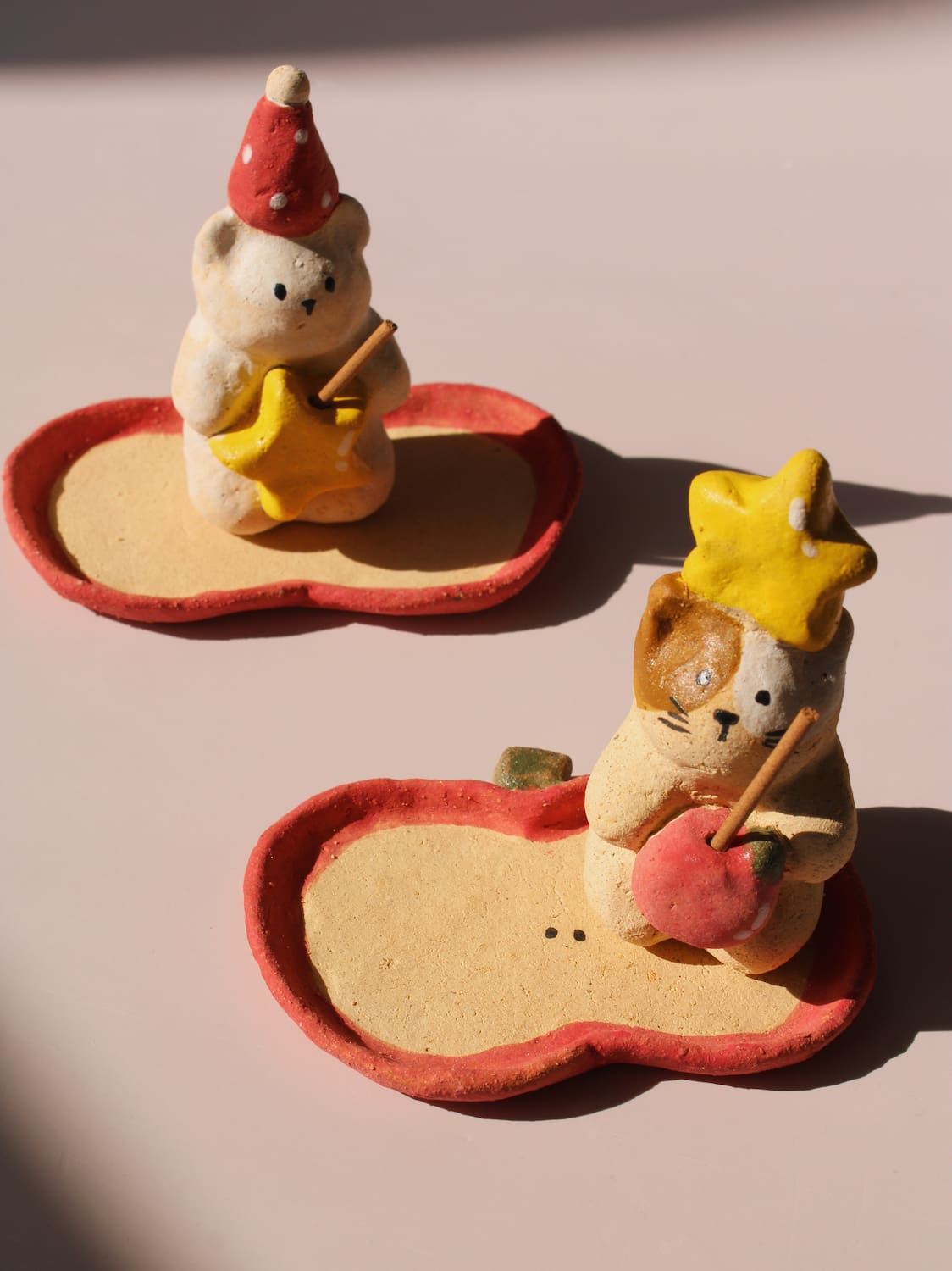 Teddy Ceramic Incense Holder and Mochi Ceramic Incense Holder, two handmade animal figurines with star and apple accents on matching apple-shaped incense trays, on a soft pink background.