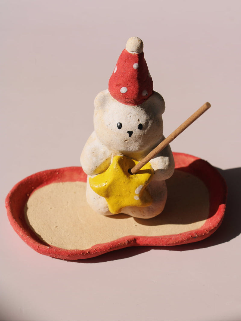 Teddy Ceramic Incense Holder, a handmade bear figurine in a red polka dot party hat holding a yellow star, paired with an apple-shaped incense tray, on a soft pink background.
