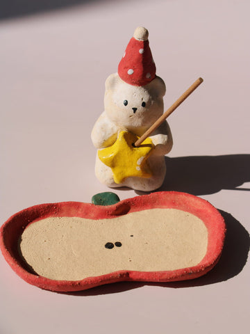 Teddy Ceramic Incense Holder, a handmade bear figurine in a red polka dot party hat holding a yellow star, paired with an apple-shaped incense tray, on a soft pink background.