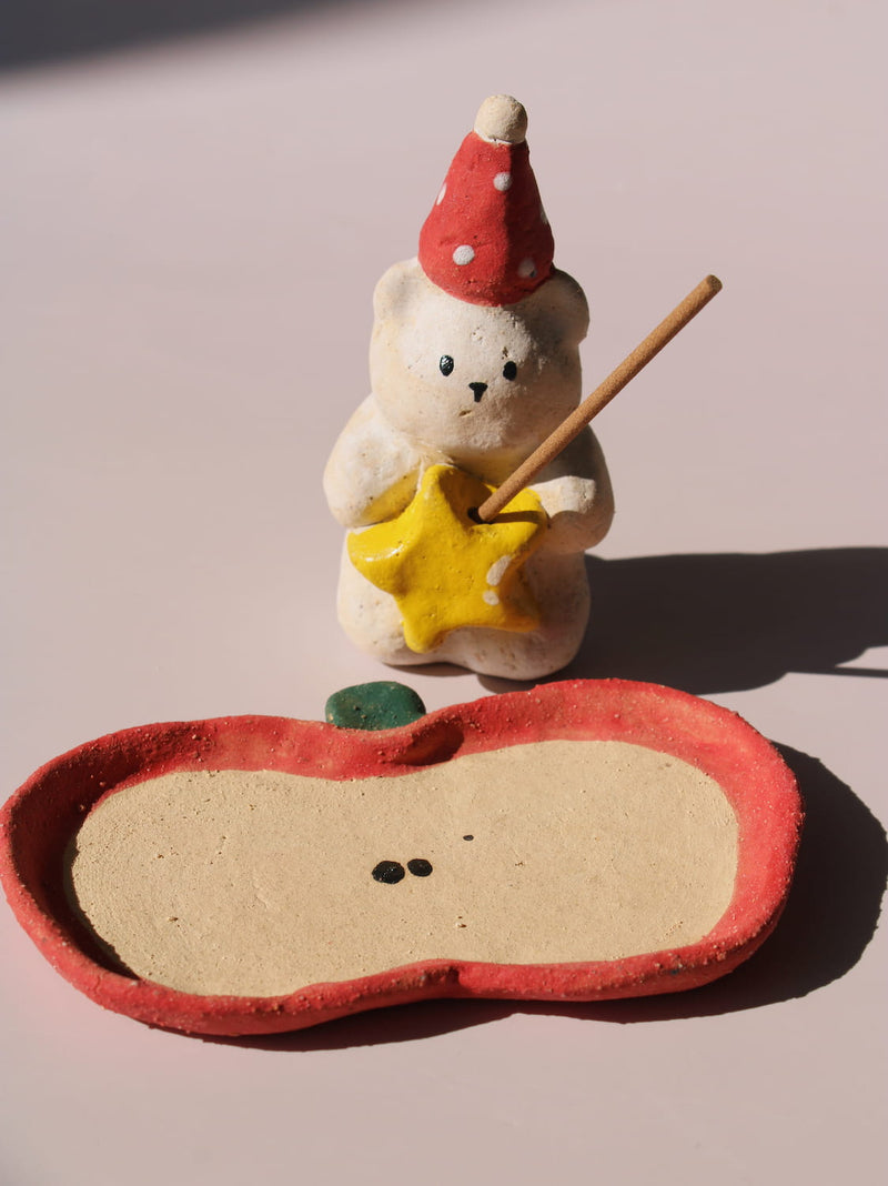 Teddy Ceramic Incense Holder, a handmade bear figurine in a red polka dot party hat holding a yellow star, paired with an apple-shaped incense tray, on a soft pink background.