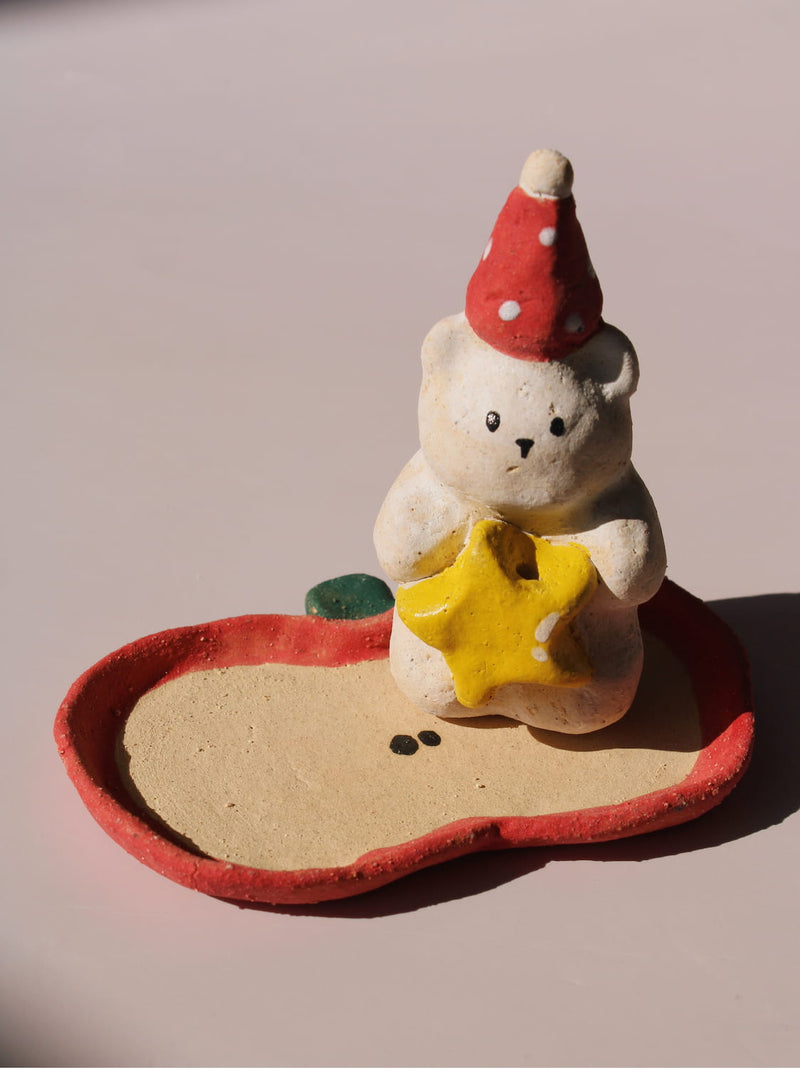 Teddy Ceramic Incense Holder, a handmade bear figurine in a red polka dot party hat holding a yellow star, paired with an apple-shaped incense tray, on a soft pink background.