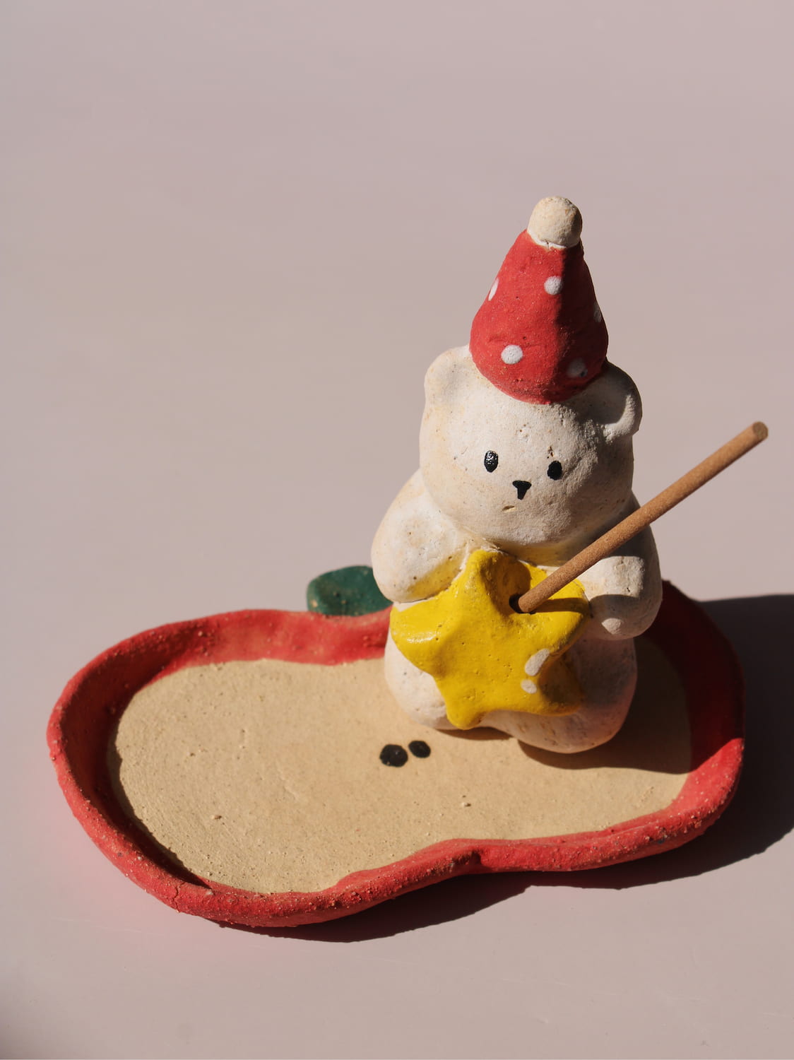 Teddy Ceramic Incense Holder, a handmade bear figurine in a red polka dot party hat holding a yellow star, paired with an apple-shaped incense tray, on a soft pink background.