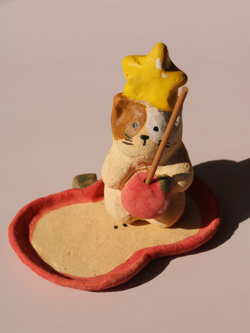 Mochi Ceramic Incense Holder, a handmade cat figurine wearing a yellow star hat and holding a pink apple, paired with an apple-shaped incense tray, on a soft pink background.