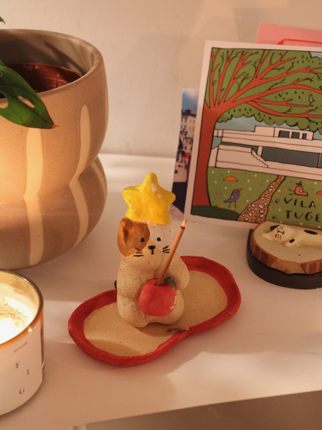 Mochi Ceramic Incense Holder with a lit incense stick, styled on a shelf with colourful artwork and a terracotta vase in the background in warm indoor light.