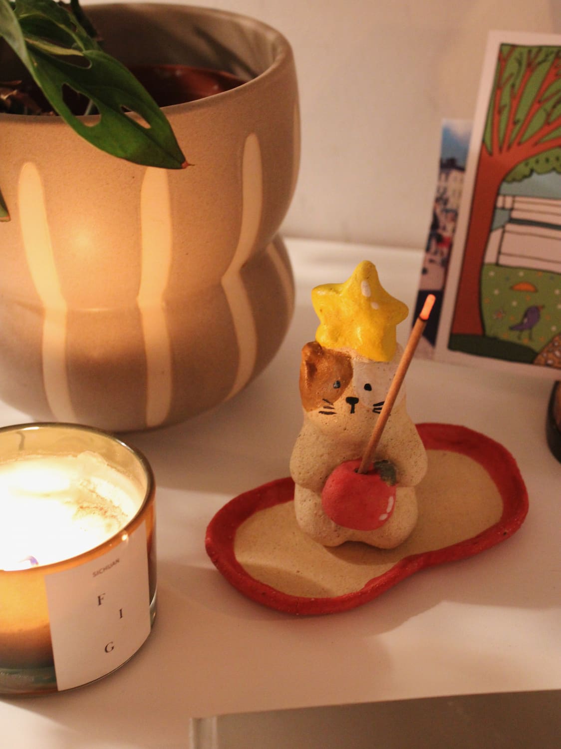 Mochi Ceramic Incense Holder with a lit incense stick, styled on a shelf with colourful artwork and a terracotta vase in the background in warm indoor light.