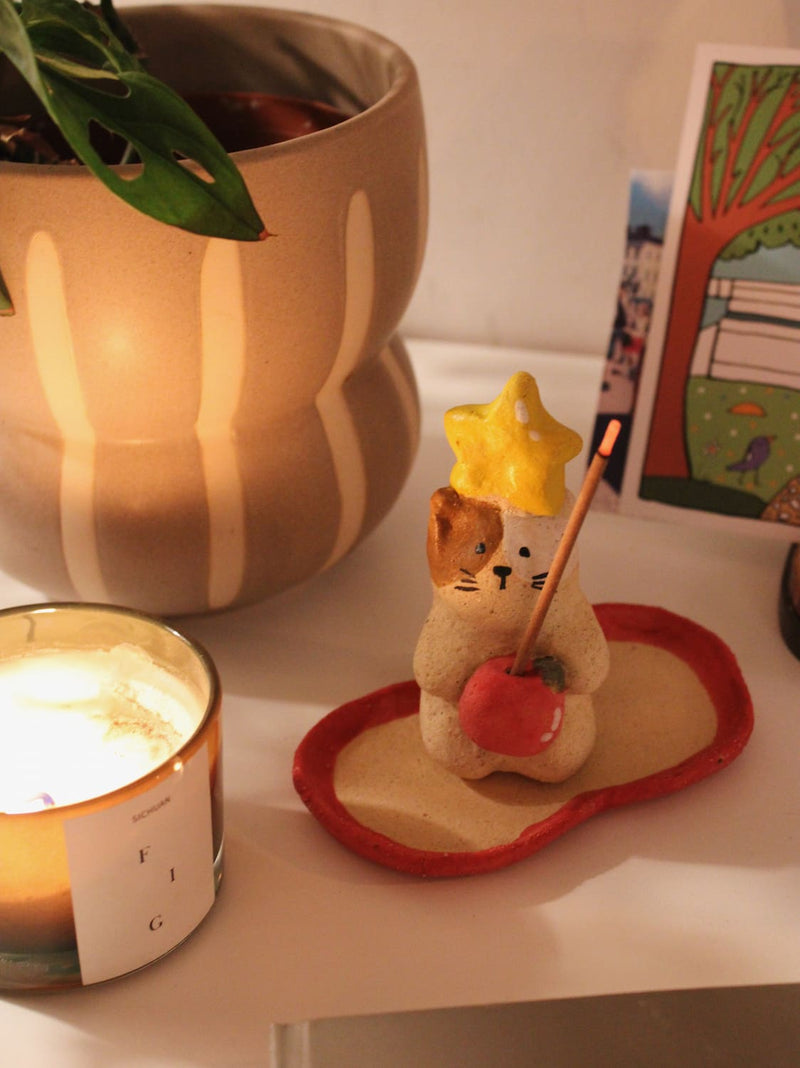 Mochi Ceramic Incense Holder with a lit incense stick, styled on a shelf with colourful artwork and a terracotta vase in the background in warm indoor light.