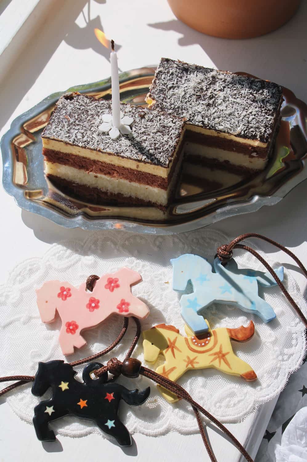 Pony Charm Necklaces in all four colorways — Petal Pony in pink with flowers, Stardust Pony in pale blue with stars, Amber Pony in yellow with western detail and Midnight Pony in black with colourful stars — displayed on a lace cloth beside a layered chocolate cake with a lit candle in natural sunlight.