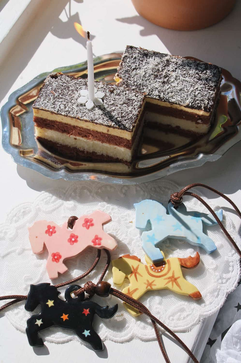 Pony Charm Necklaces in all four colorways — Petal Pony in pink with flowers, Stardust Pony in pale blue with stars, Amber Pony in yellow with western detail and Midnight Pony in black with colourful stars — displayed on a lace cloth beside a layered chocolate cake with a lit candle in natural sunlight.