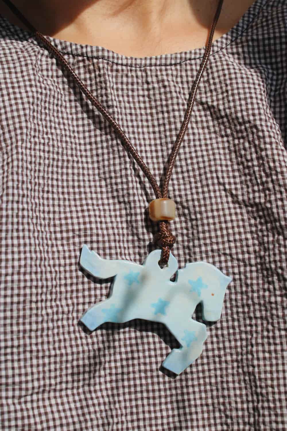 Stardust Pony Charm Necklace in pale blue with star detail worn on a brown gingham top in natural sunlight.